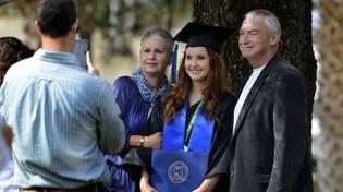 女毕业生戴着帽子，穿着长袍，拿着文凭，和父母合影.